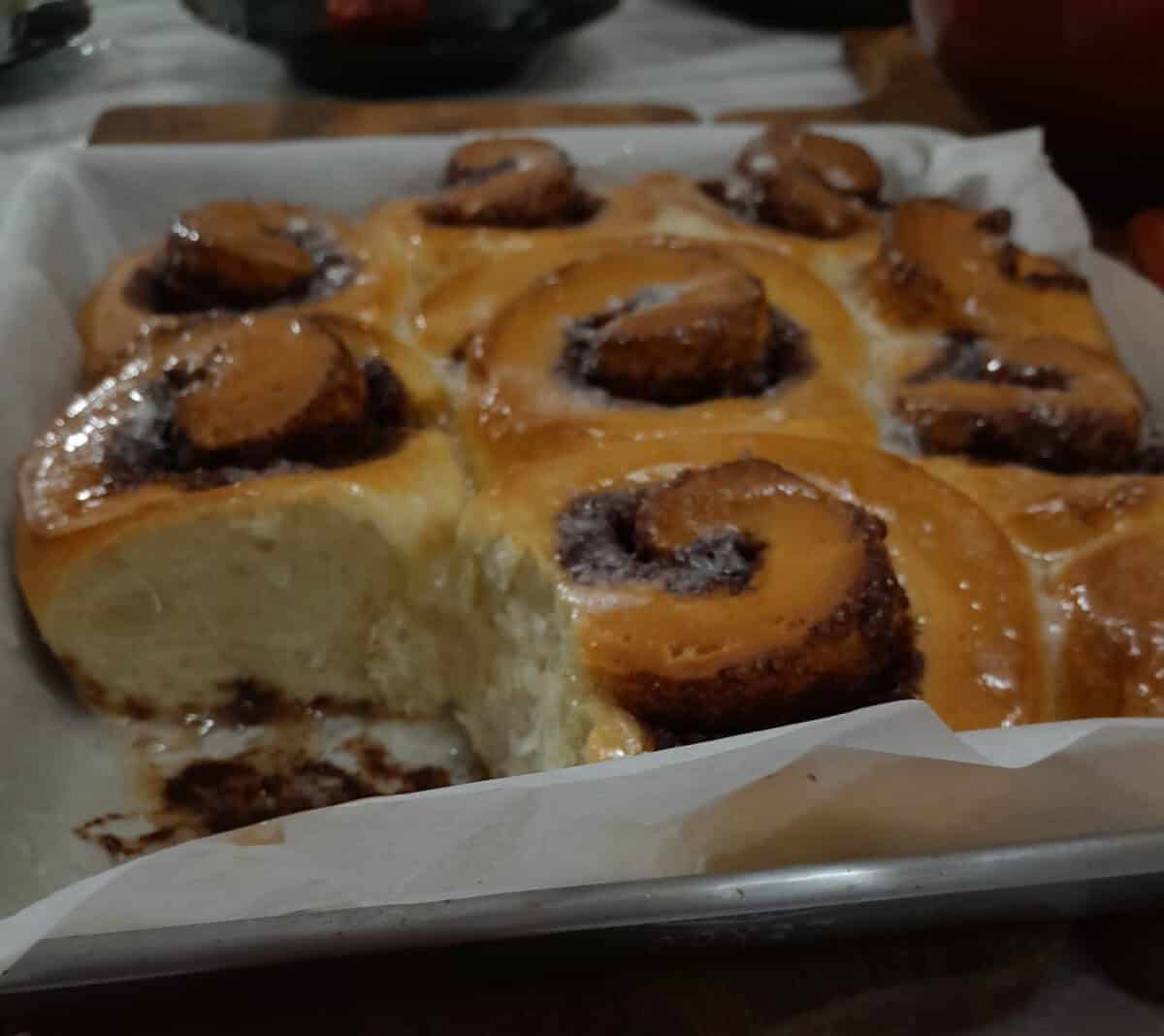 A tray of freshly baked cinnamon rolls, some with a shiny glaze on top. One roll has been taken out, revealing the soft, fluffy interior and swirls of cinnamon filling.