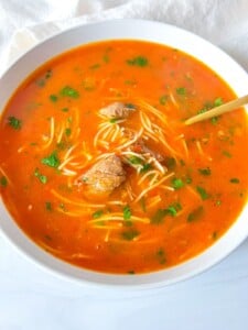 A bowl of red broth beef soup with thin noodles, tender chunks of meat, and fresh chopped herbs, served with a spoon—an inviting Beef Soup Recipe to warm your day.