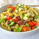 A bowl of the best pasta salad featuring rotini, cherry tomatoes, cucumbers, mozzarella balls, Kalamata olives, sun-dried tomatoes, and fresh basil, all tossed in a light dressing.