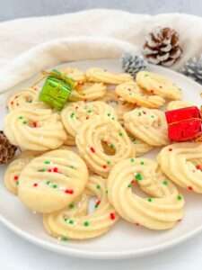 A plate of Shortbread Butter Cookies topped with red, white, and green sprinkles, surrounded by small holiday gift boxes and pinecones on a white surface.
