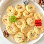 A white plate filled with Shortbread Butter Cookies topped with red and green sprinkles, surrounded by pinecones and small wrapped gifts, creates a festive holiday display.