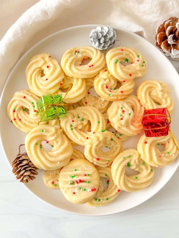 A white plate filled with Shortbread Butter Cookies topped with red and green sprinkles, surrounded by pinecones and small wrapped gifts, creates a festive holiday display.