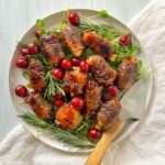 A white plate with grilled bacon-wrapped chicken pieces and bacon wrapped dates, garnished with fresh rosemary, green sprouts, and bright red cherries, with a serving spoon on the side and a white napkin underneath.