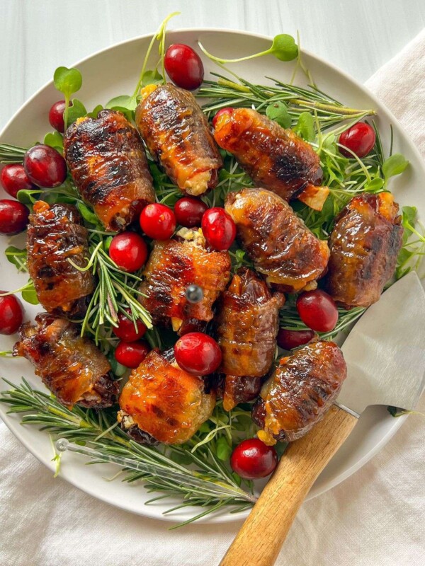 A white plate with grilled bacon-wrapped chicken pieces and bacon wrapped dates, garnished with fresh rosemary, green sprouts, and bright red cherries, with a serving spoon on the side and a white napkin underneath.