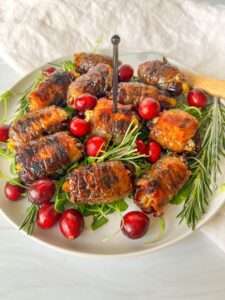 A plate of Bacon Wrapped Dates arranged on fresh greens, garnished with sprigs of rosemary and whole cranberries, with a pick in the center for easy serving.
