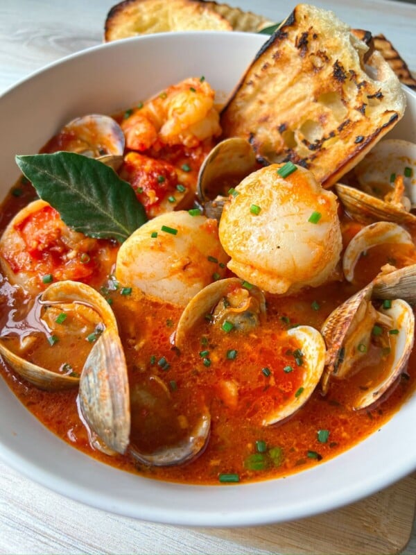 A bowl of Cioppino Recipe seafood stew with shrimp, scallops, clams, and a rich red tomato broth, garnished with chives and a bay leaf, served alongside toasted bread slices.