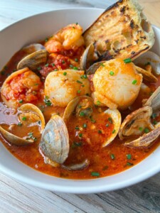 A bowl of seafood stew inspired by a classic Cioppino recipe, filled with shrimp, scallops, clams, and tomato-based broth, garnished with chopped chives and served with a slice of toasted bread.