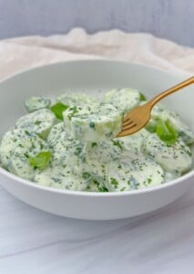 A white bowl filled with creamy, herbed Polish Cucumber Salad, garnished with fresh basil leaves. A gold fork holds up a round cucumber slice coated in thick, green-speckled dressing.