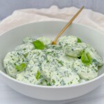 A white bowl filled with Creamy Cucumber Salad—sliced cucumbers coated in a rich herb dressing and garnished with fresh green herbs—rests on a light surface with a gold fork inside.