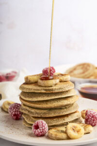 A stack of pancakes topped with banana slices and raspberries on a white plate. Syrup is being poured over the top. In the background, there are more pancakes and a small bowl of syrup.