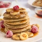 A stack of healthy banana pancakes topped with banana slices and raspberries, drizzled with syrup, on a white plate. Surrounding the pancakes are additional banana slices and raspberries. A fork and small bowl of syrup are in the background.