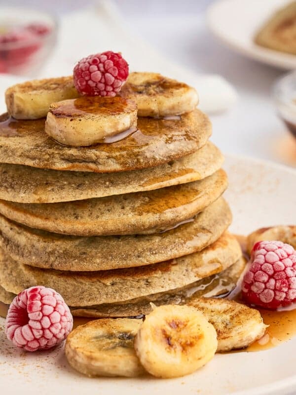 A stack of healthy banana pancakes topped with banana slices and raspberries, drizzled with syrup, on a white plate. Surrounding the pancakes are additional banana slices and raspberries. A fork and small bowl of syrup are in the background.