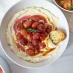 A white bowl filled with creamy hummus and whipped feta dip, topped with roasted grapes, a drizzle of sauce, and a sprig of thyme. A toasted bread slice rests on the edge, with a plate of more toasted bread nearby.
