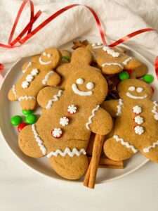 A plate of Easy Gingerbread Cookies shaped like people, adorned with white icing details, red and green candy buttons, cinnamon sticks, and colorful candies, set on a white surface with a red ribbon and white cloth.