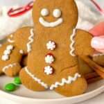 A hand holds an Easy Gingerbread Cookie decorated with white icing and red and white candy buttons, shaped like a smiling gingerbread person. More cookies and green candy are on the plate in the background.