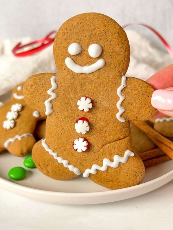 A hand holds an Easy Gingerbread Cookie decorated with white icing and red and white candy buttons, shaped like a smiling gingerbread person. More cookies and green candy are on the plate in the background.