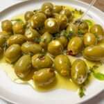 A plate of feta cheese olives, drizzled with olive oil and garnished with fresh thyme and red pepper flakes. A clear fork rests on the plate.