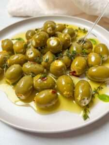 A plate of feta cheese olives, drizzled with olive oil and garnished with fresh thyme and red pepper flakes. A clear fork rests on the plate.