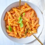 A bowl of Penne alla Vodka coated in creamy tomato sauce, garnished with fresh basil leaves and herbs, with two gold forks resting in the bowl. A white cloth is placed beside the bowl.