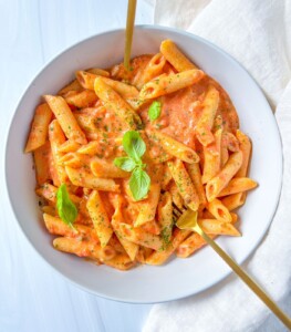 A bowl of Penne alla Vodka coated in creamy tomato sauce, garnished with fresh basil leaves and herbs, with two gold forks resting in the bowl. A white cloth is placed beside the bowl.