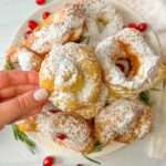 A hand holds a powdered sugar-covered zeppole above a plate piled with similar pastries, garnished with cranberries and rosemary, on a white surface.