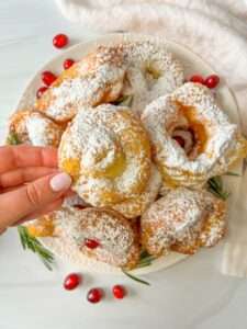 A hand holds a powdered sugar-covered zeppole above a plate piled with similar pastries, garnished with cranberries and rosemary, on a white surface.