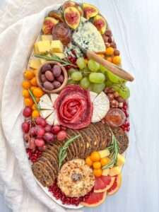 A Holiday Cheese Board featuring various cheeses, salami roses, fresh figs, grapes, cherry tomatoes, olives, crackers, grapefruit slices, rosemary sprigs, and bowls of jam is artfully arranged on a white surface with a linen cloth.