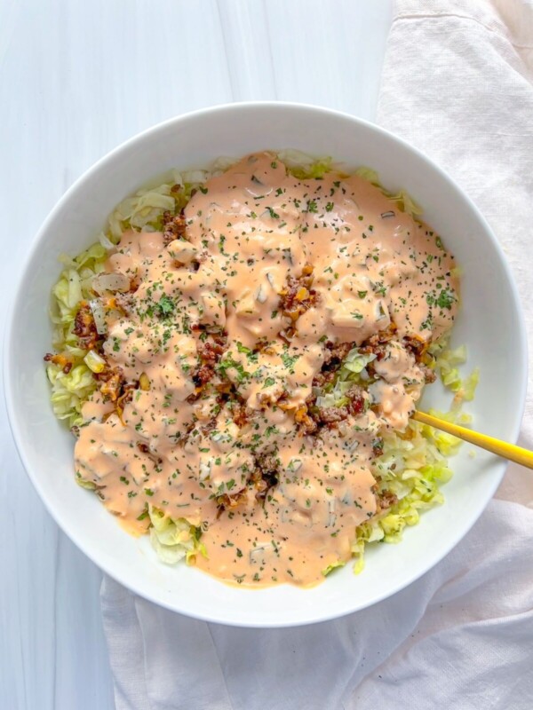 A white bowl filled with chopped lettuce, ground beef, and a generous layer of creamy orange Big Mac Salad sauce, garnished with herbs. A yellow fork rests in the bowl, which sits on a white surface next to a light napkin.