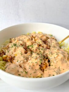 A white bowl filled with rice, ground meat, and corn, all topped with a creamy orange sauce and garnished with chopped herbs sits beside a gold-colored spoon—a deconstructed take on a Mason Jar Salad.