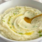 A bowl of creamy mashed potatoes garnished with chopped herbs and a drizzle of melted butter, served alongside whole roasted cauliflower, with a gold spoon resting in the mash.