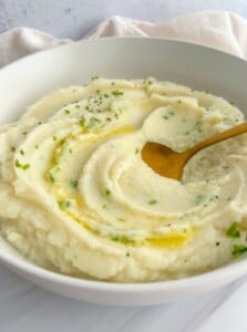 A bowl of creamy mashed potatoes garnished with chopped herbs and a drizzle of melted butter, served alongside whole roasted cauliflower, with a gold spoon resting in the mash.