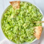A bowl of chopped green salad with lettuce and cucumber, dressed lightly and topped with three triangular pita chips, sits on a white surface with a white napkin nearby—perfect alongside dishes using fresh or dried herbs from your How to Dry Herbs guide.