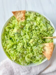 A bowl of chopped green salad with lettuce and cucumber, dressed lightly and topped with three triangular pita chips, sits on a white surface with a white napkin nearby—perfect alongside dishes using fresh or dried herbs from your How to Dry Herbs guide.