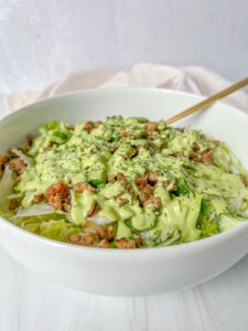 A white bowl filled with a green salad topped with crumbled meat and drizzled with a creamy green dressing—perfect for using fresh greens after learning how to dry herbs—rests beside a fork ready for your first bite.