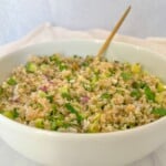 A white bowl filled with a fresh tabbouleh salad, inspired by the Jennifer Aniston Salad, featuring bulgur, chopped parsley, green vegetables, red onions, and a spoon, set against a light background.