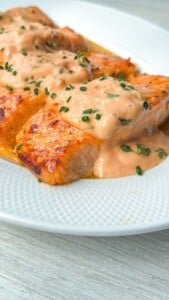 Mac Sauce Salmon fillets are grilled to perfection, topped with a creamy, pink sauce and garnished with chopped herbs, served on a white, textured plate.