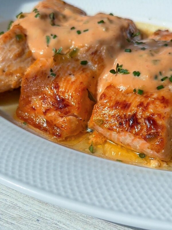 Close-up of Mac Sauce Salmon: three pieces of grilled salmon topped with a creamy orange sauce and fresh herbs, served on a white plate.