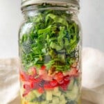 A vibrant Mason Jar Salad, this glass jar is filled with layered diced cucumbers, tomatoes, bell peppers, walnuts, green onions, and spinach atop yellow vinaigrette, all sealed fresh with a metal lid.