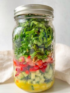A vibrant Mason Jar Salad, this glass jar is filled with layered diced cucumbers, tomatoes, bell peppers, walnuts, green onions, and spinach atop yellow vinaigrette, all sealed fresh with a metal lid.