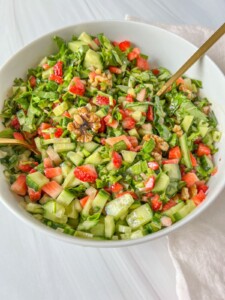 A white bowl filled with a fresh Jennifer Aniston Salad made of chopped cucumbers, strawberries, greens, and walnuts, with a gold spoon and napkin set on a light surface.