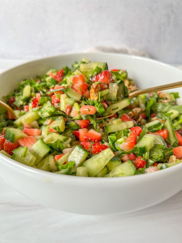 A white bowl filled with Strawberry Spinach Salad made of chopped cucumbers, strawberries, leafy greens, and walnuts, with a fork resting inside.