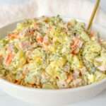 A white bowl filled with creamy Olivier Salad, featuring diced potatoes, carrots, pickles, ham, and herbs, with a gold spoon resting inside.