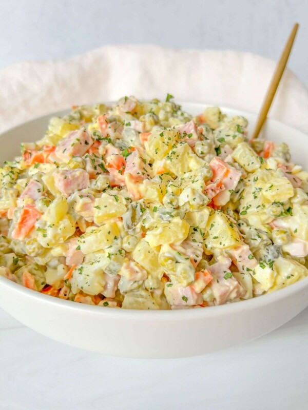 A white bowl filled with creamy Olivier Salad, featuring diced potatoes, carrots, pickles, ham, and herbs, with a gold spoon resting inside.