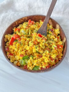 A wooden bowl filled with colorful tofu scramble, made using an easy tofu scramble recipe with chopped red and yellow bell peppers, green onions, and herbs. A wooden spoon rests inside. The bowl sits on a white surface with a white cloth nearby.