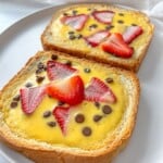 Two slices of bread topped with a creamy custard, fresh strawberry slices, and chocolate chips are served on a white plate. The background is light and softly blurred.