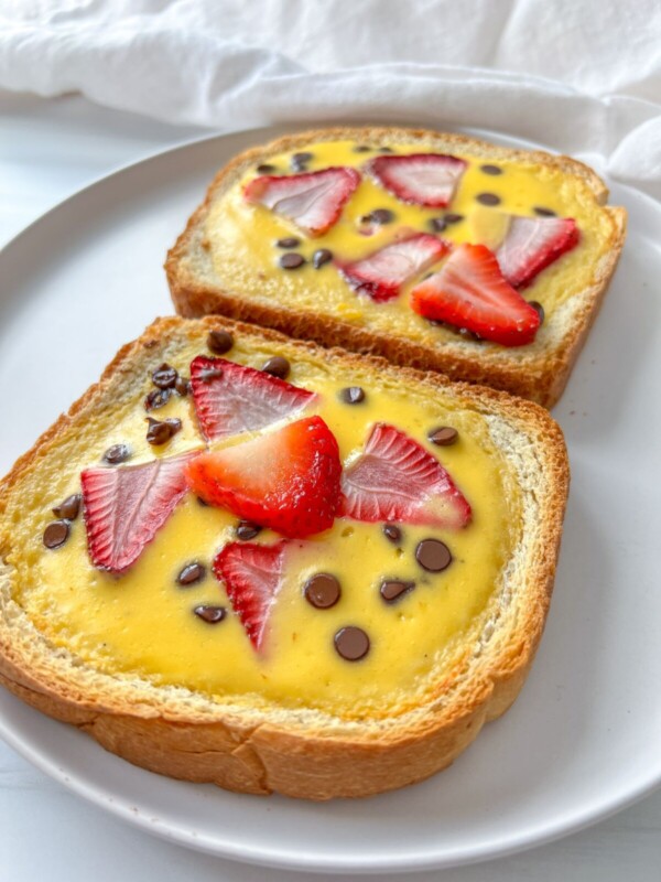 Two slices of bread topped with a creamy custard, fresh strawberry slices, and chocolate chips are served on a white plate. The background is light and softly blurred.