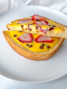 Two slices of bread topped with yellow custard, fresh strawberry slices, and chocolate chips are stacked on a white plate.
