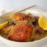 A bowl of glazed Garlic Lemon Chicken Thighs garnished with herbs, served with lemon wedges and a gold fork resting on the edge.