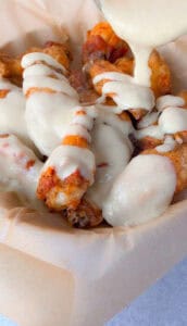 Alfredo Chicken Wings in a parchment-lined tray are being topped with a generous drizzle of creamy white sauce.