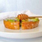 A slice of toasted bread topped with creamy avocado and drizzled honey sits on a white plate—a simple presentation inspired by the Best Avocado Toast Recipe, with a honey dipper resting on top.